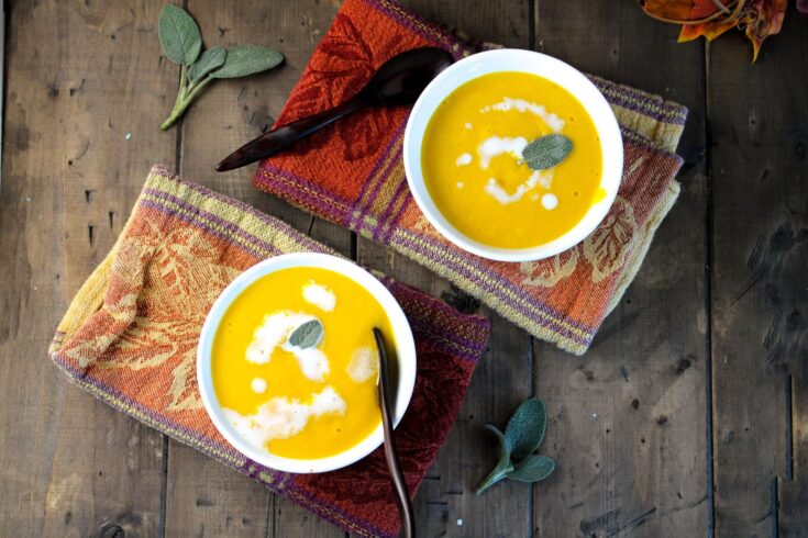 Creamy pumpkin soup garnished with sage leaves served in white bowls on colorful woven napkins, set on a rustic wooden table for a cozy, autumn-inspired meal.