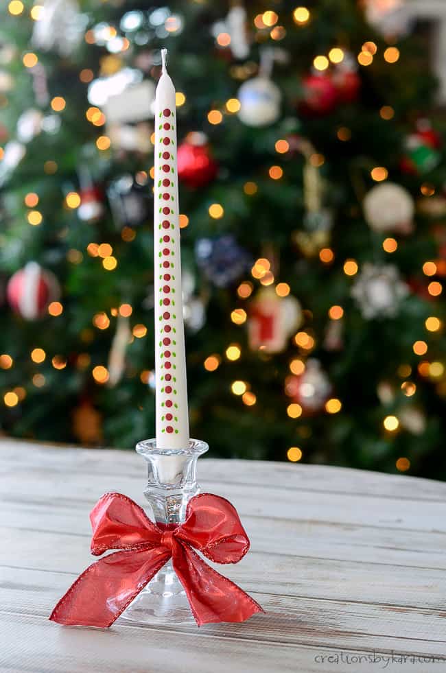 Pink polka dot candle in a glass holder with a red ribbon bow, set against a decorated Christmas tree with colorful ornaments and warm lights, perfect for holiday decor.