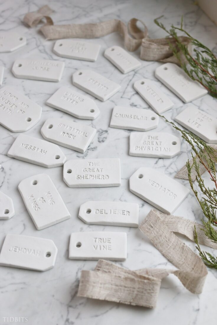 Fragile white marble church tags with engraved faith-inspired words like "Messiah," "Ying of Kisses," and "Counselor" scattered on a marble surface, decorative ribbons and greenery.