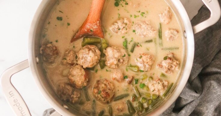 Savory homemade meatball soup in a creamy broth with green beans and herbs, cooked in a stainless steel pot for comforting meal.