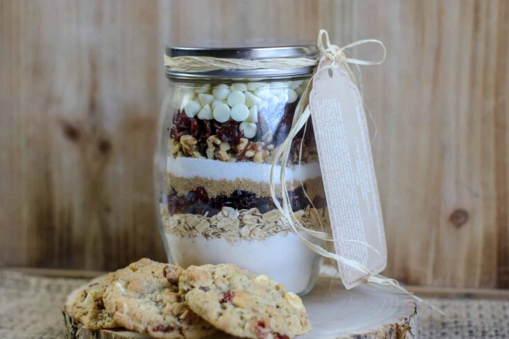 Layers of cookie crumbs, dried fruit, white chocolate, chopped nuts, sugar, and other ingredients in a transparent jar, with a cookie in front against a wooden background.