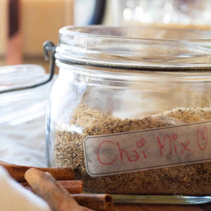 Chai spice mix in a glass jar labeled "Chai Mix" with cinnamon sticks nearby, showcasing ingredients used in homemade chai tea preparation.
