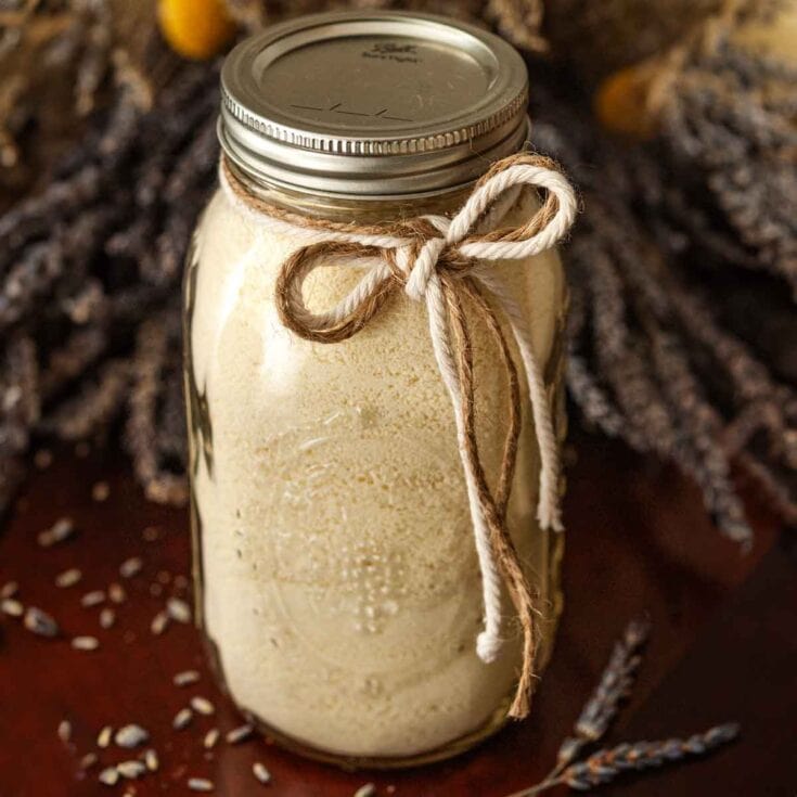 Creamy homemade vanilla body scrub in glass jar with rustic twine bow, surrounded by dried lavender and sesame seeds, emphasizing natural skincare and self-care routines.