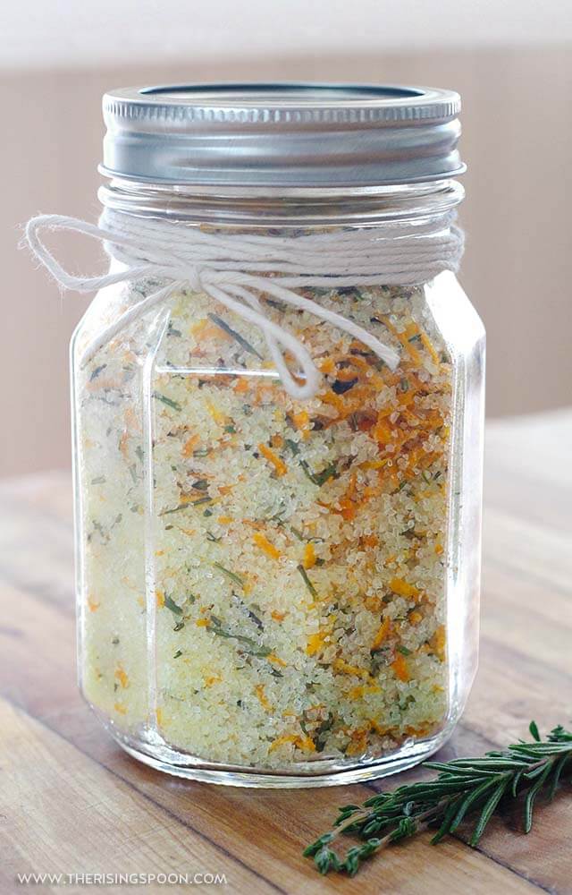Flavored bath salt in a glass jar with a silver lid and twine tie, featuring colorful herbs and flowers for relaxation.