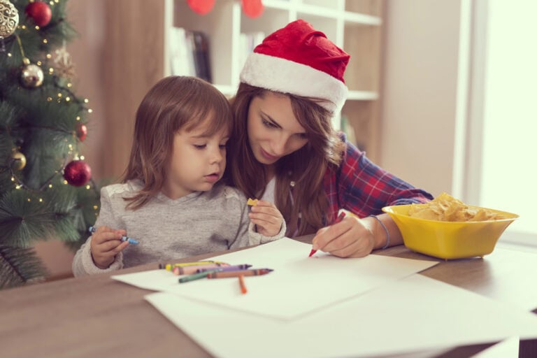 Festive holiday scene featuring a young woman in a Santa hat and a little girl drawing and coloring at a wooden table, surrounded by Christmas decorations and a bowl of snacks, emphasizing warmth and family Christmas traditions.