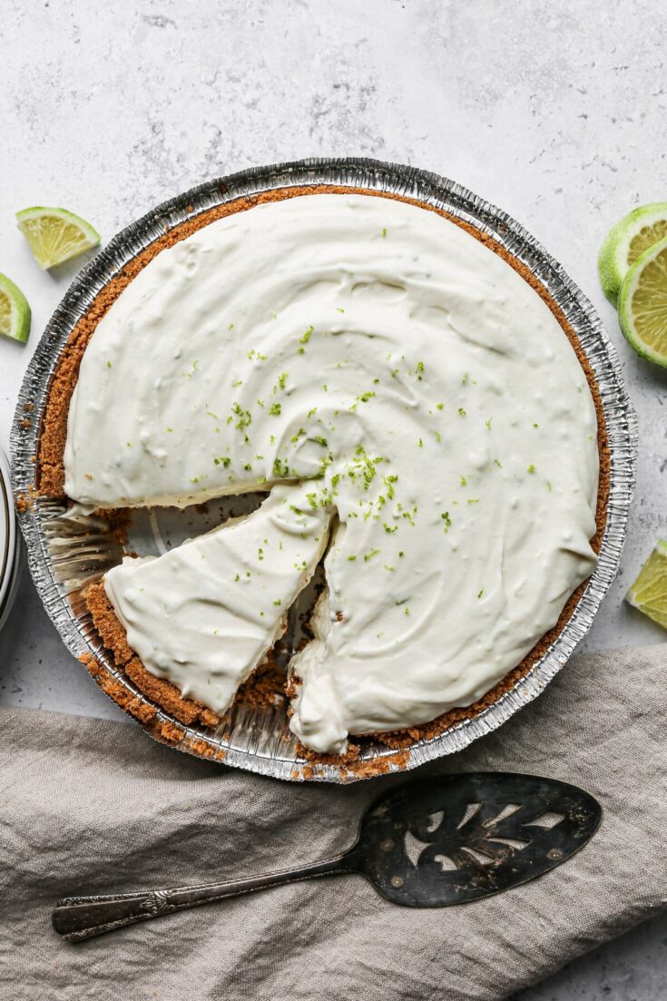 Creamy lime cheesecake with a graham cracker crust, topped with lime zest, served in a disposable aluminum pan, garnished with fresh lime wedges and a vintage cake server.