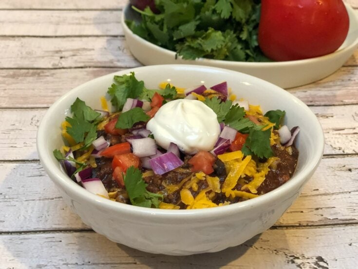 Savory chili topped with shredded cheese, diced tomatoes, red onions, fresh cilantro, and a dollop of sour cream in a white bowl, on a rustic wooden table, showcasing comforting meal options from A Hundred Affections.