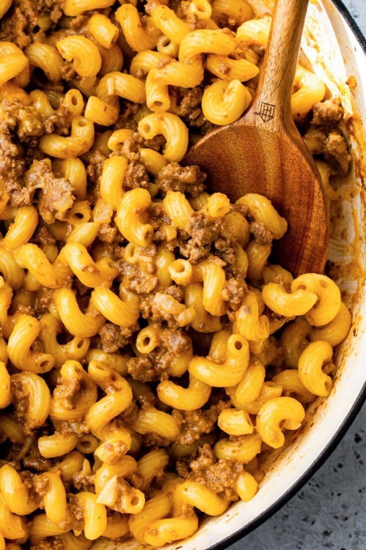 Creamy baked macaroni and cheese with beef ground meat in a white casserole dish with a wooden spoon.