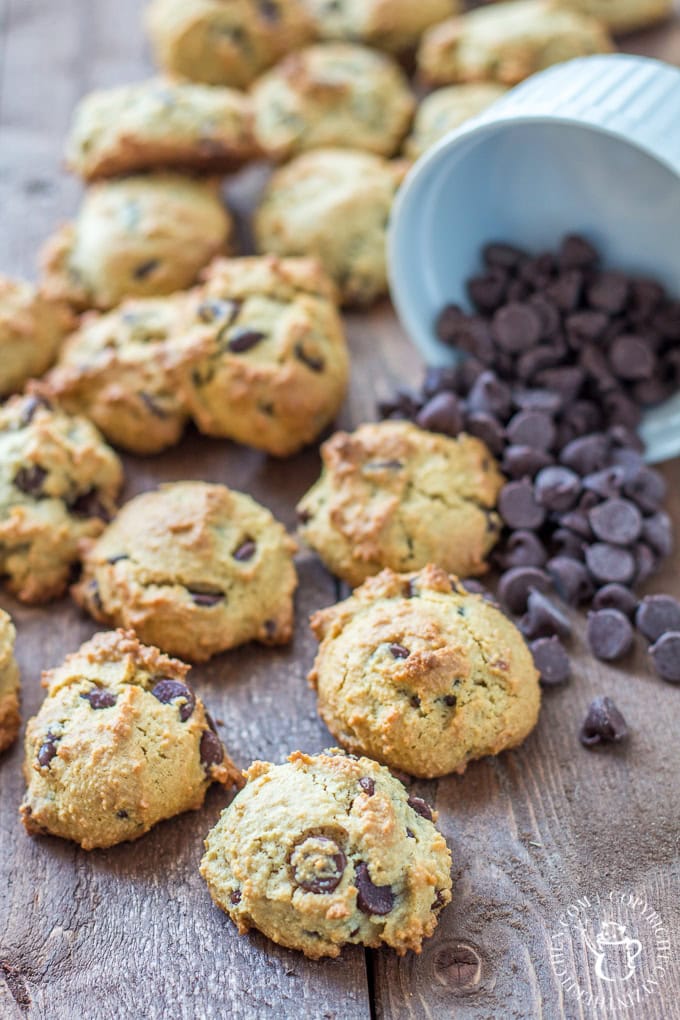 Soft homemade chocolate chip cookies with a golden crust, scattered on a rustic wooden surface. A tipped-over blue mug spills dark chocolate chips, adding a fresh, inviting touch to this classic cookie setup.