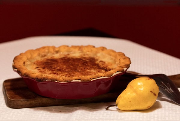 Golden baked apple pie in a red dish with a fresh pear, served on a wooden board, perfect for cozy dessert moments at A Hundred Affections.