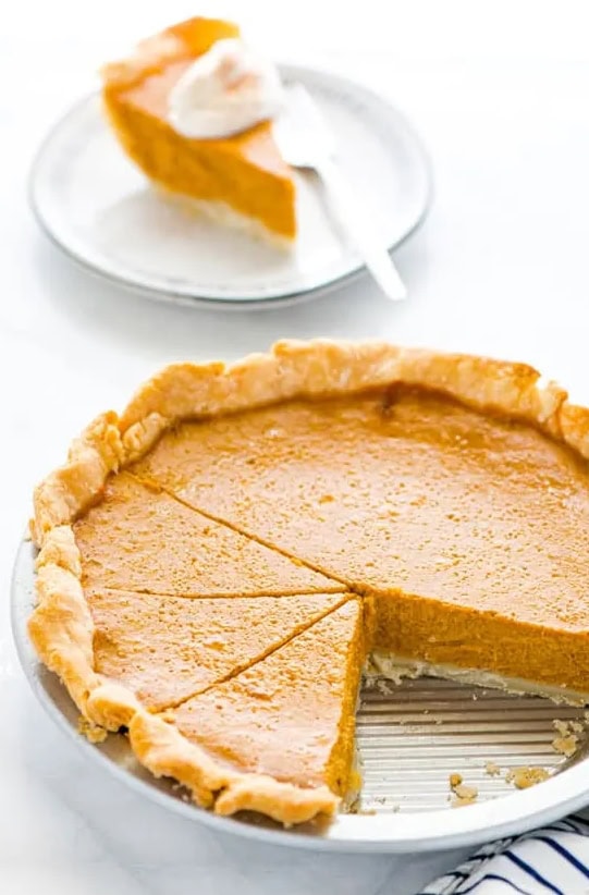 Golden pumpkin pie with a flaky crust, served on a white plate with a slice cut out, and a slice of pumpkin pie with whipped cream on a separate dish, showcasing classic fall desserts and homemade Thanksgiving treats.