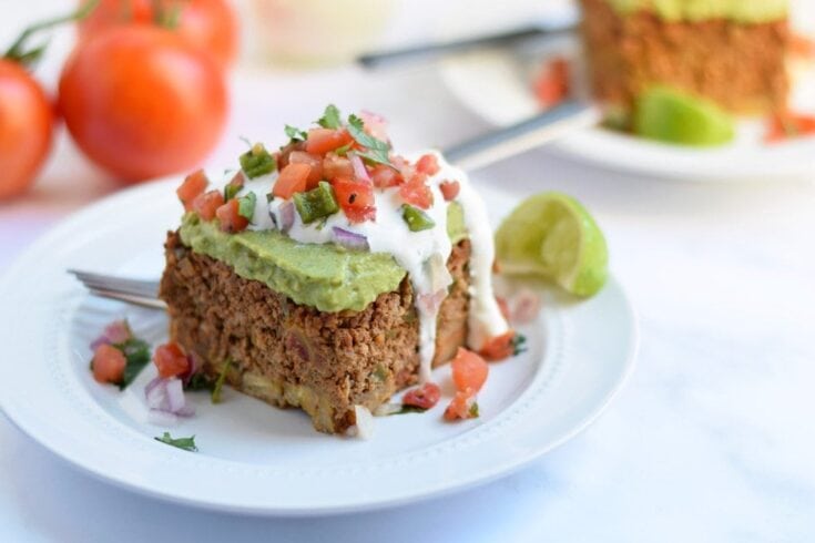 Freshly baked Taco Pie topped with guacamole, sour cream, and pico de gallo, served on a white plate with lime wedges, highlighting flavorful Mexican comfort food from A Hundred Affections.