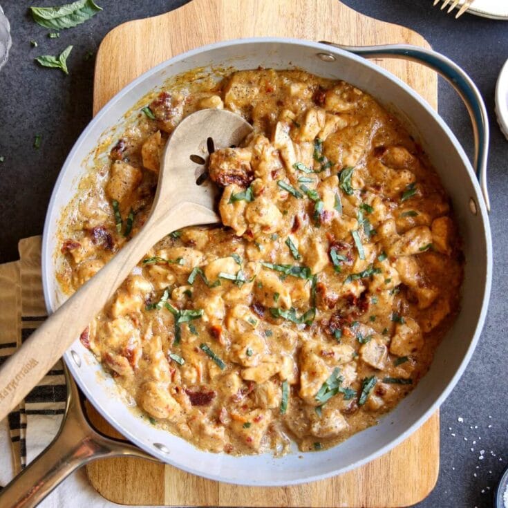 Creamy chicken stew with sun-dried tomatoes and fresh herbs in a white skillet, served on a wooden cutting board, highlighting comfort food and homestyle cooking on a cozy table setting.