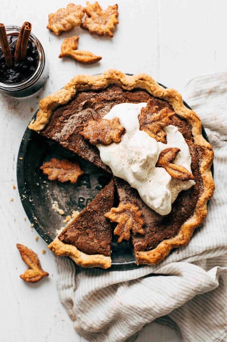 Rich pumpkin pie with whipped cream and leaf-shaped crust decorations, served on a black plate with cinnamon sticks and a jar of dark syrup, perfect for fall desserts.