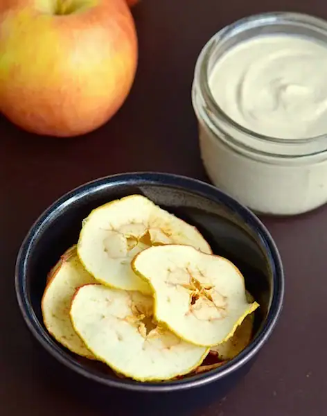 Fresh apple slices in a black bowl with natural yogurt or cream in a glass jar, ripe apple in the background, healthy snack or dessert concept.