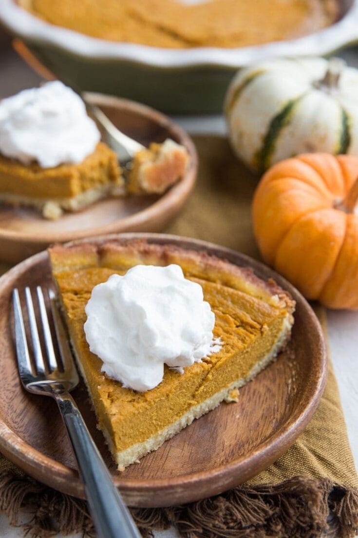 Pumpkin pie with whipped cream served on a rustic wooden plate, surrounded by small decorative pumpkins, autumn-themed setting, perfect for fall dessert recipes.