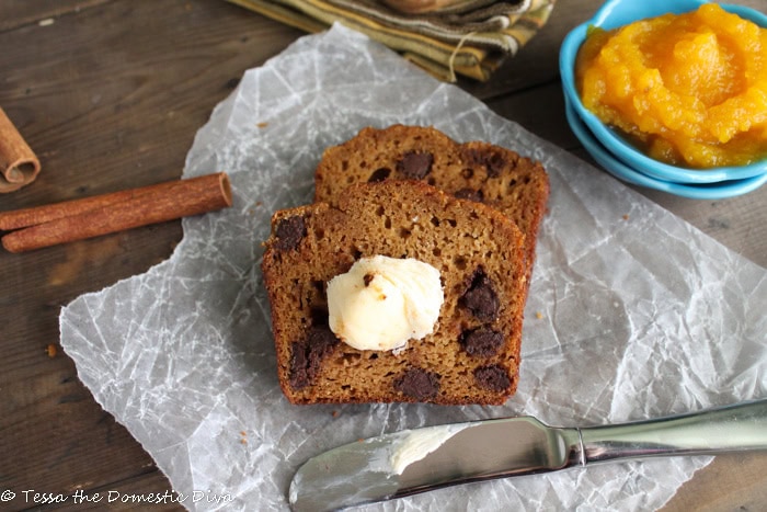 Chocolate chip banana bread slices topped with melted butter, served on crinkled parchment paper with a bowl of fruit compote and cinnamon sticks in the background, creating a cozy and inviting breakfast or snack scene.