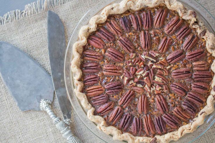 Fresh pecan pie topped with whole pecans in a clear glass dish, set on a textured fabric surface with a pie server nearby.