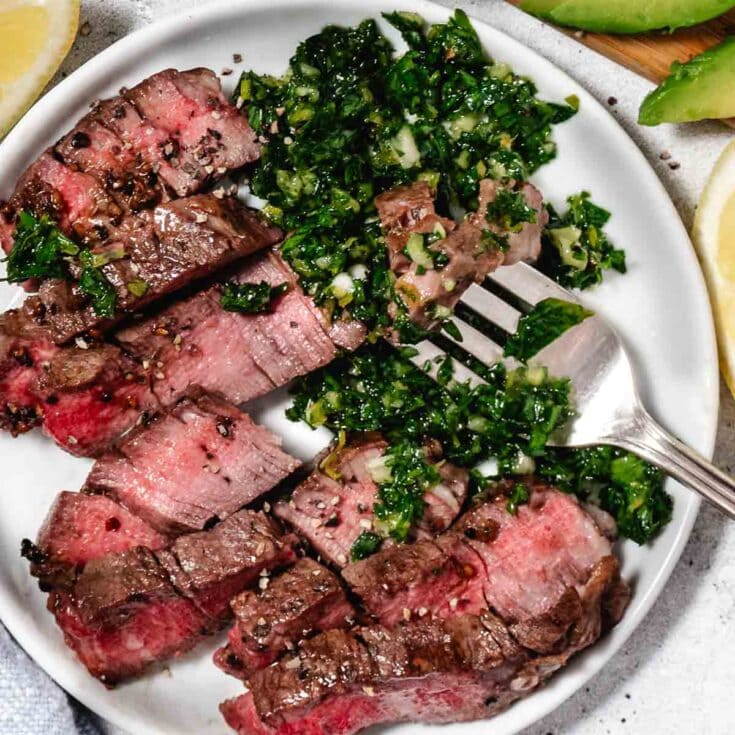 Flank steak with chimichurri sauce on a white plate serving as a healthy, flavorful meat dish for dinner or special occasions.