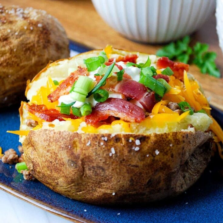 Roasted baked potato stuffed with bacon, shredded cheese, green onions, and sour cream served on a blue plate with a bread roll and a bowl in the background.