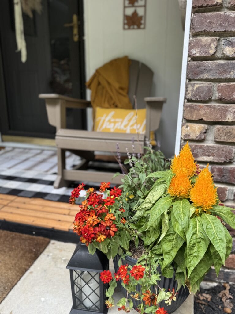 Vibrant autumn flower arrangement with red, orange, and yellow flowers in a black pot, displayed outdoors against a brick wall, 