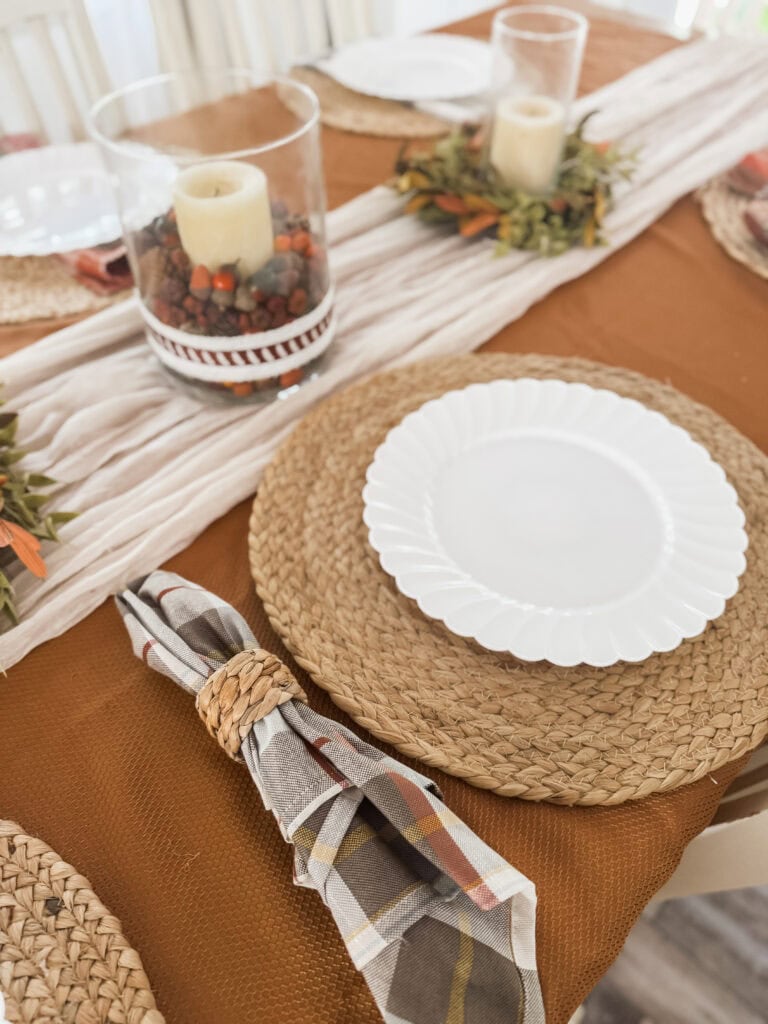 A beautifully set autumn-inspired dining table with woven placemats, a white scalloped plate, plaid napkin with a woven napkin ring, and seasonal candle centerpiece.