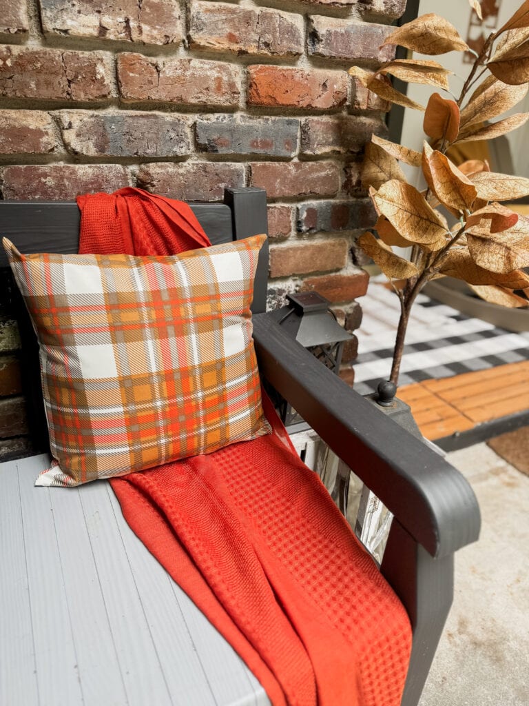 Cozy outdoor seating with plaid pillow, red blanket, and autumn decor against a rustic brick wall at A Hundred Affections.