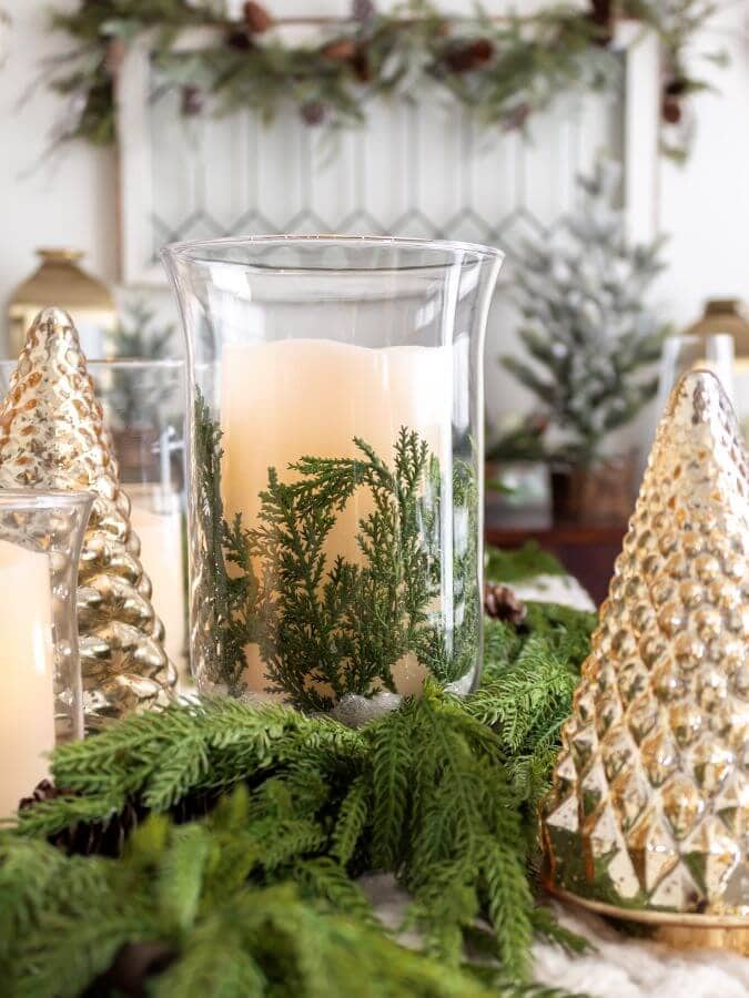Bright winter holiday table centerpiece featuring a tall glass candle holder with greenery, gold and silver decorative trees, and lush evergreen foliage, perfect for Christmas decor.