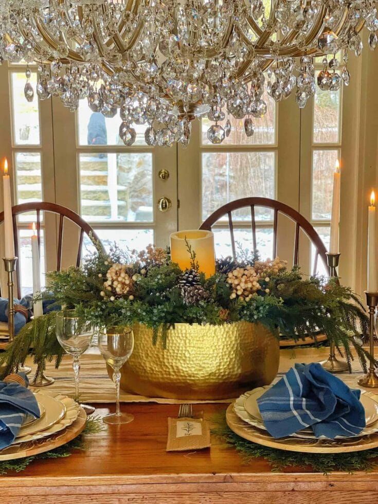 Elegant holiday dining table featuring a gold hammered centerpiece, lush green foliage, white berries, pinecones, and candle accents under a crystal chandelier.