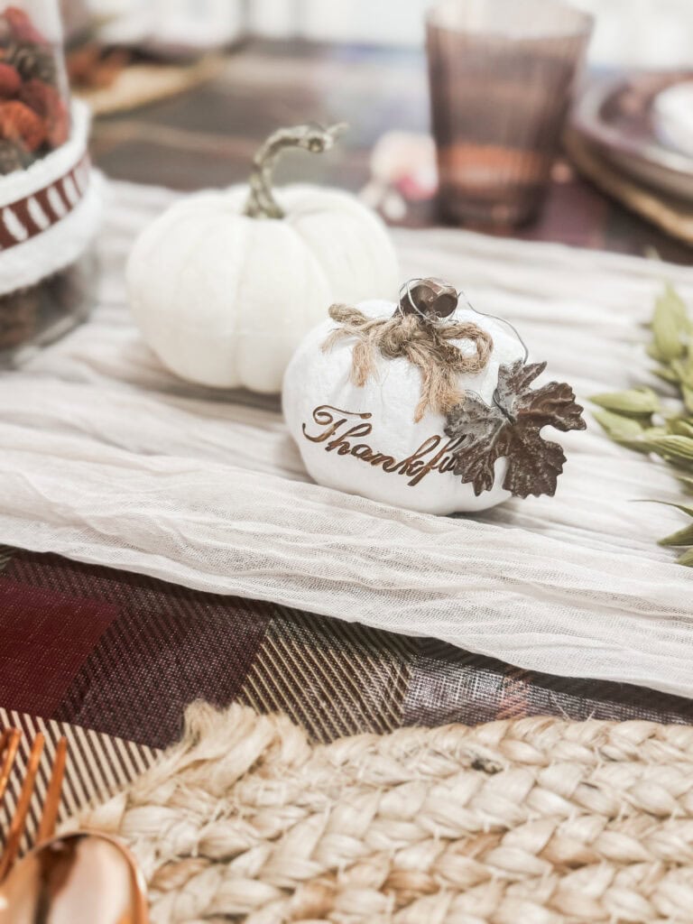 White pumpkin with "Thankful" message and fall leaf decorations on a rustic table setting.