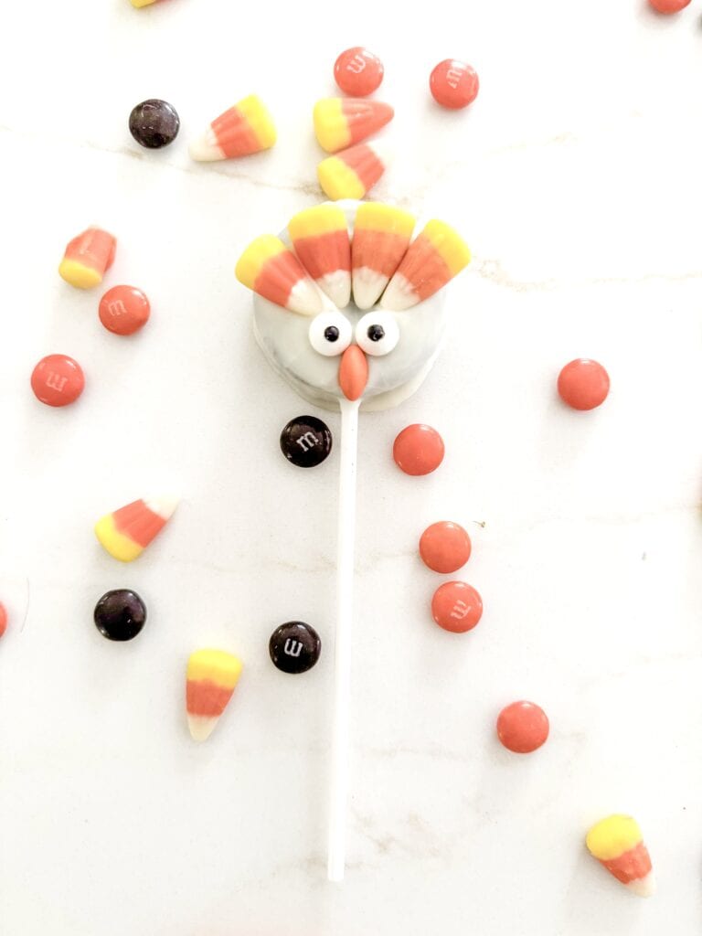 Colorful turkey-shaped candy pop with googly eyes and candy corn feathers, surrounded by assorted M&M's and candy pieces on white background.