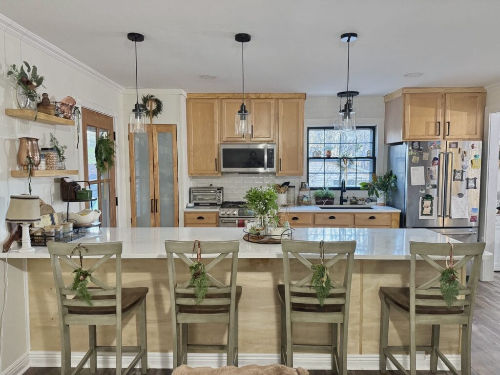 Cozy farmhouse kitchen with natural wood cabinets, white marble island, and festive green wreaths on chairs, ideal for holiday gatherings and homey decor.