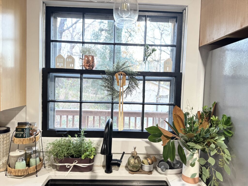 Bright kitchen window with potted plants, decorative vases, and cozy decor, featuring a black window frame, natural wood accents, and a view of trees outside.