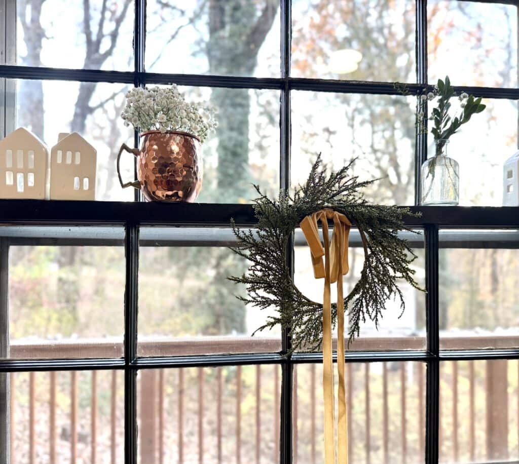 Copper mug with white flowers and decorative house-shaped ornaments on window sill, cozy holiday decor in a bright home, perfect for Christmas interior styling.