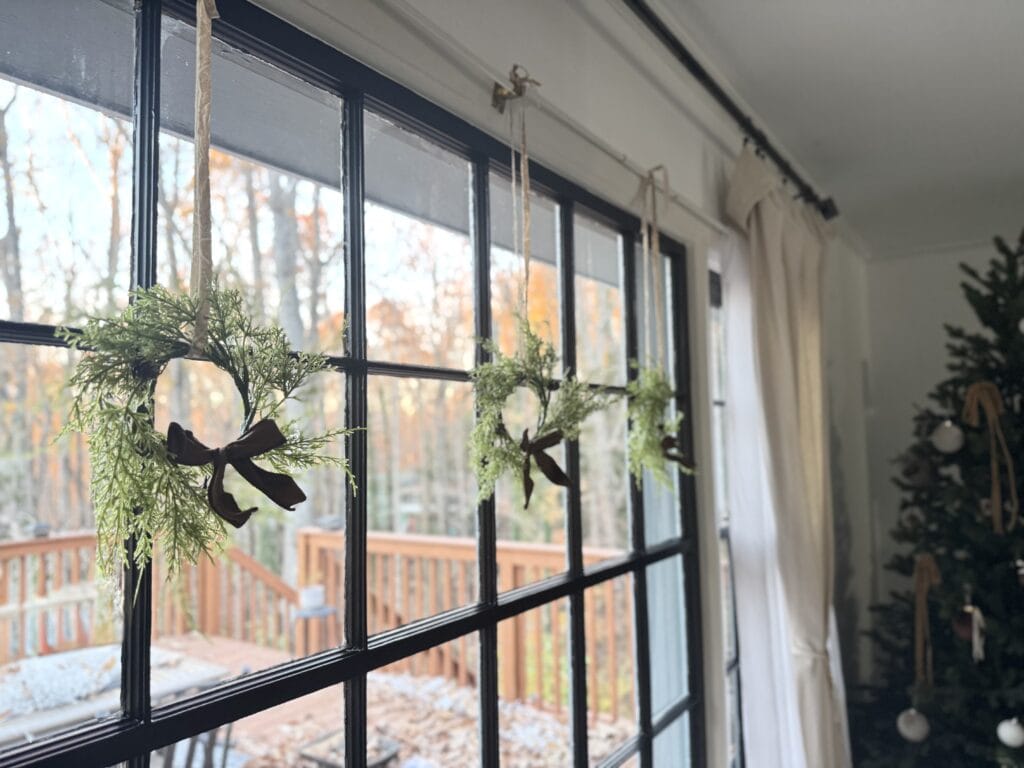 Festive holiday decorations hanging on a large window with a cozy, decorated Christmas tree in the background, creating a warm, inviting holiday atmosphere.