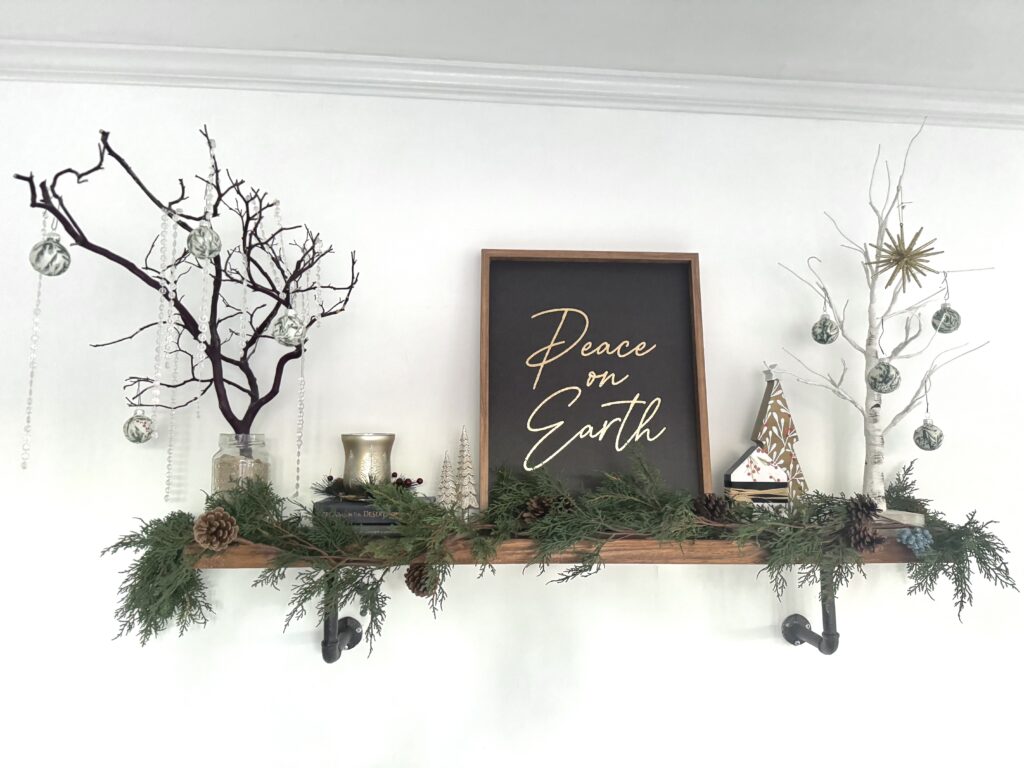 Rustic holiday decor with white and dark branches, Christmas ornaments, greenery, and a "Peace on Earth" framed sign on a wooden shelf.