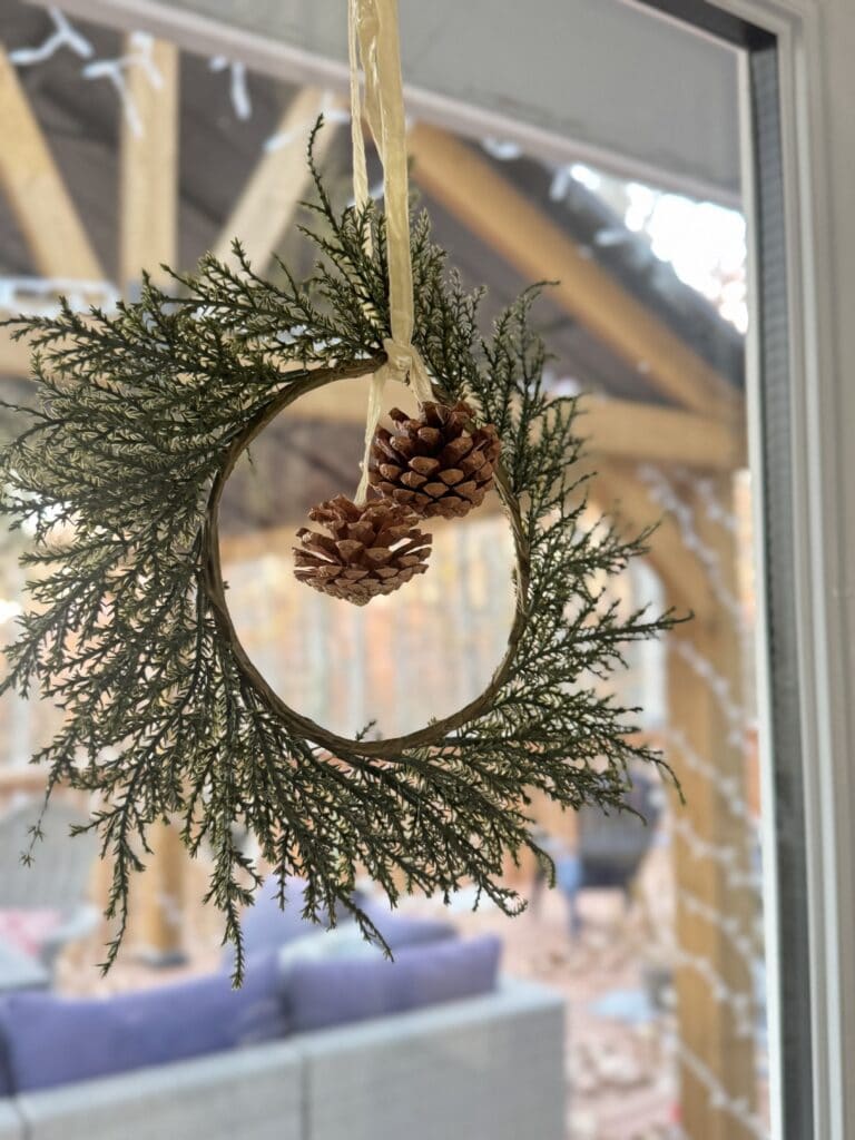 Handmade Christmas wreath hanging inside, decorated with pinecones and greenery, on a window with blurred outdoor background.