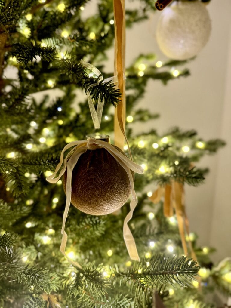 Golden Christmas ornament with cream ribbons hanging on a festive decorated tree, warm glowing lights, and elegant holiday decor.
