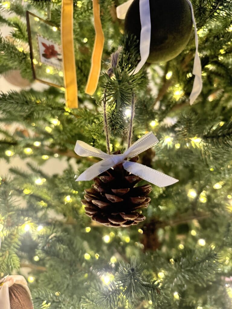 Pinecone Christmas ornament with white ribbon hanging on a decorated holiday tree, warm lights in background.