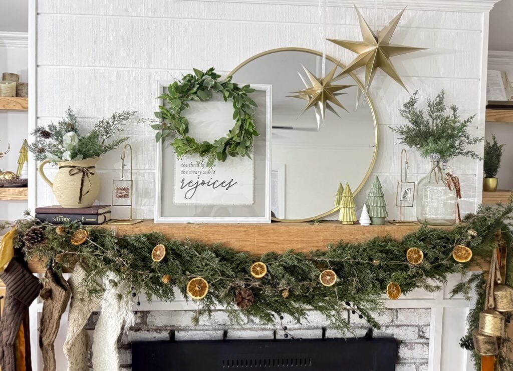Festive holiday mantel decorated with greenery, dried orange slices, candles, framed quote, gold star ornaments, and small decorative trees in a cozy Christmas setting.