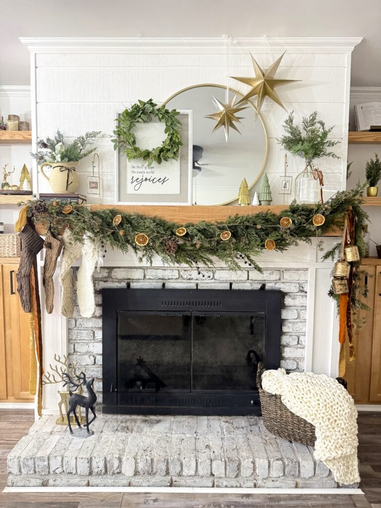 Festive holiday Christmas mantel decor with green garland, dried orange slices, stockings, and decorative ornaments in a cozy living room setting.