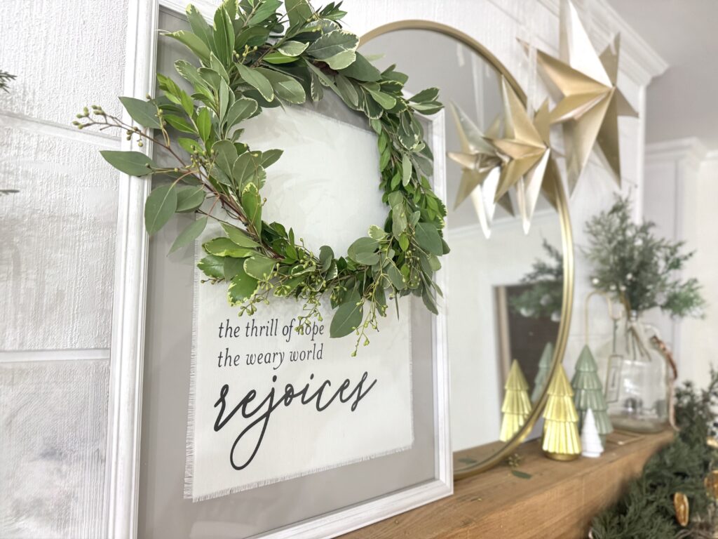 Lush holiday greenery wreath with variegated leaves and small berries, framed with white, hanging above a decorative sign with inspiring message, on rustic wooden mantel.
