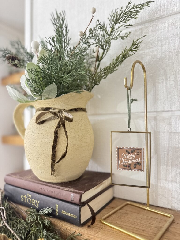 Festive holiday arrangement with a creamy textured vase, greenery, and a vintage-style "Merry Christmas" sign on a gold stand. Perfect for Christmas home decor and seasonal decorations.