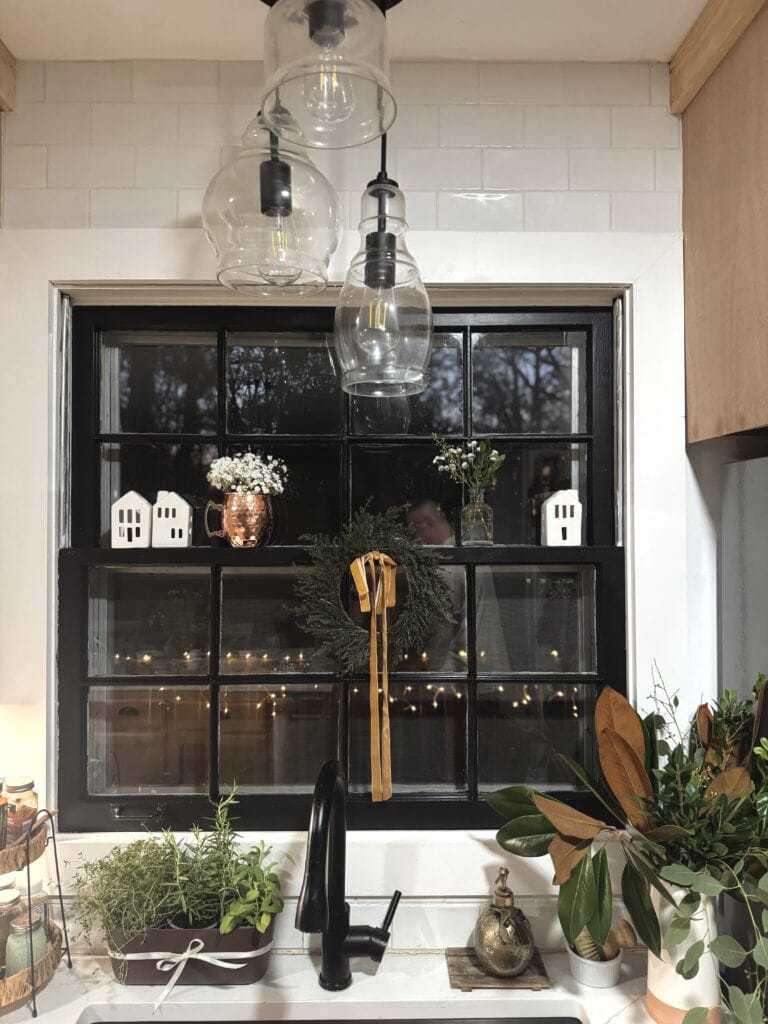 Rustic black-framed kitchen window with holiday wreath, potted herbs and greenery, cozy decor, and warm lighting for seasonal home ambiance.