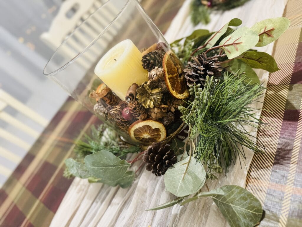 Festive holiday centerpiece with pinecones, dried orange slices, greenery, and candle in a glass holder, perfect for Christmas or winter decor.