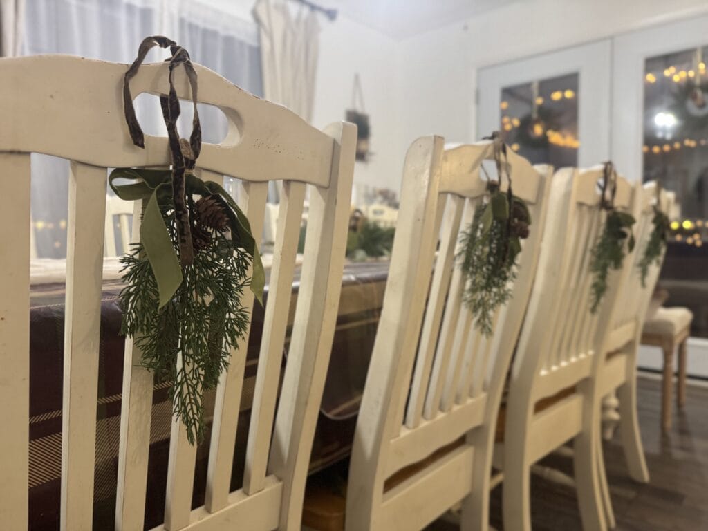 Delightful holiday table decor with white wooden chairs decorated with fresh greenery and pinecones, perfect for winter gatherings or Christmas celebrations.