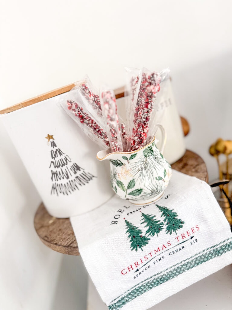 Assorted peppermint candy canes in a decorative teapot on a holiday-themed cloth with Christmas trees.