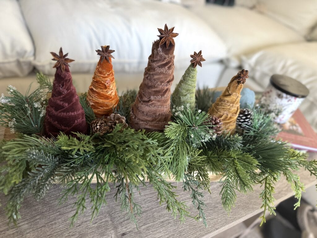 Colorful fabric Christmas tree decorations with star toppers on greenery and pinecones arrangement for holiday decor.