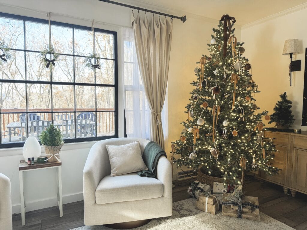 Festively decorated Christmas tree with ornaments and wrapped gifts in a cozy living room with large window and armchair.