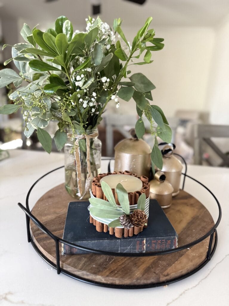 Vase with greenery and white flowers on a round wooden tray with candles, surrounded by decorative elements, perfect for rustic home decor and natural-inspired table settings.